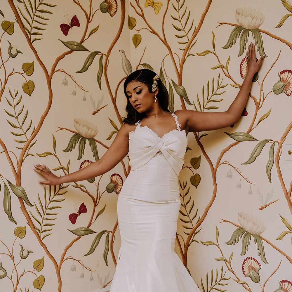 Bride wearing bow front sleeveless satin wedding dress with large gold bridal hair clip posing at Braxted Park wedding venue with floral wallpaper 