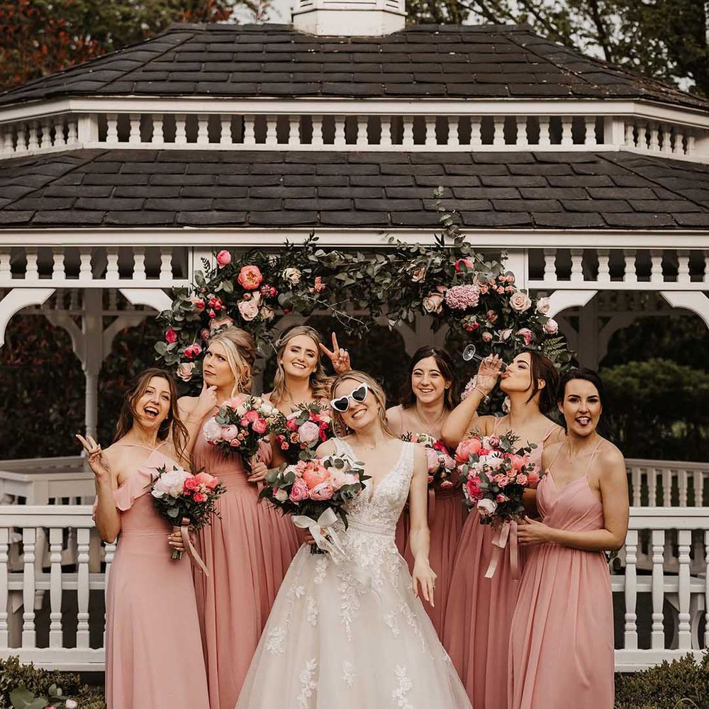 Bride in 3d applique embellished wedding dress standing by pink flower arch in wedding sunglasses with the bridal party in sleeveless pink bridesmaid dresses 