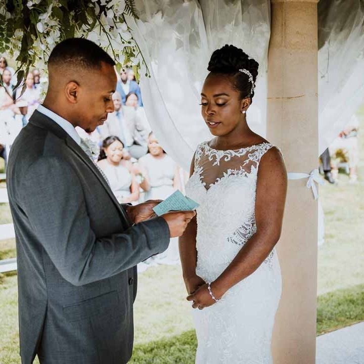 Groom in charcoal grey grooms suit, light grey patterned waistcoat, light grey patterned tie and large white rose and foliage boutonniere reading vows to bride in lace and mesh boatneck wedding dress with silver bridal hair accessories at Tredudwell Manor wedding 