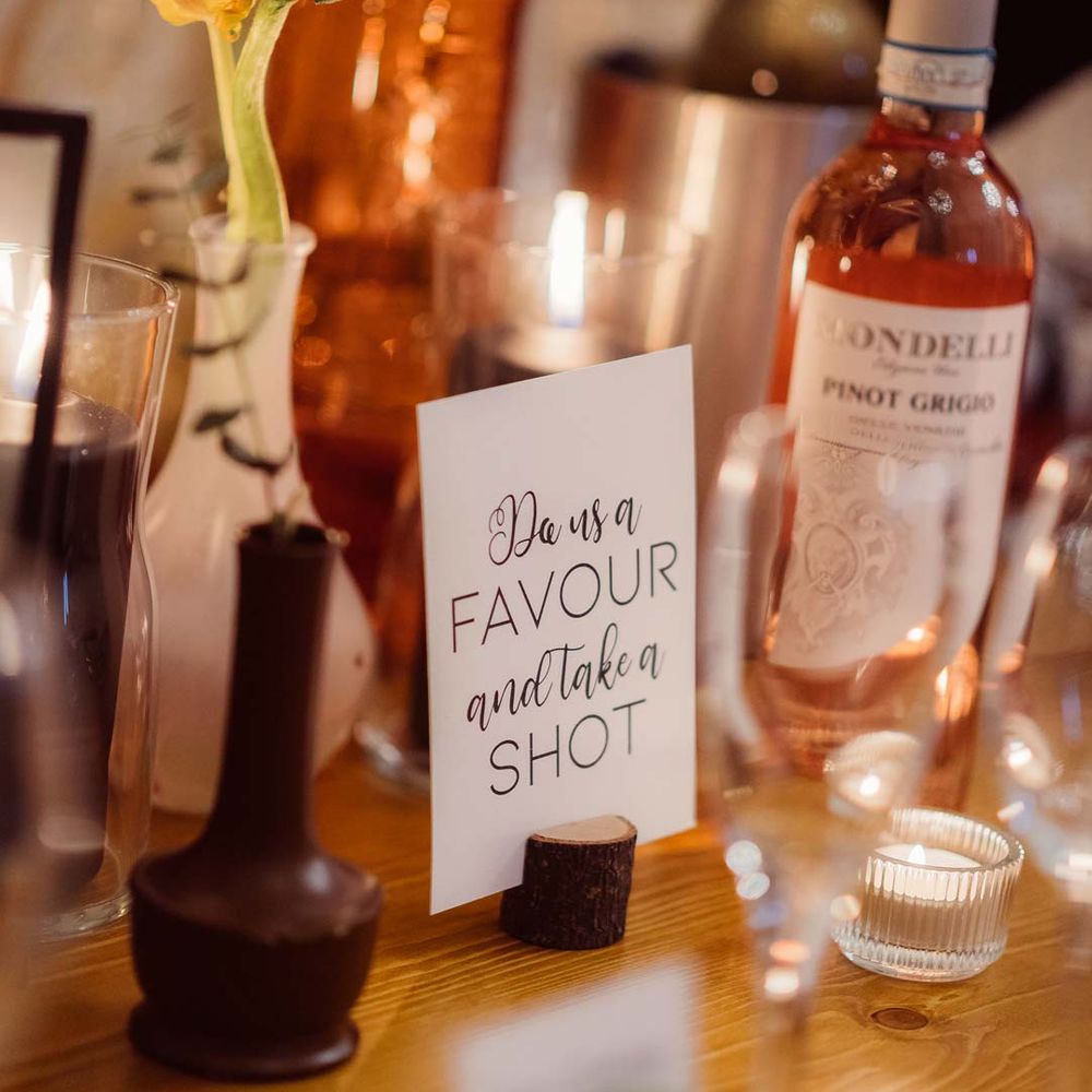 Minimalist white wedding sign "Do us a favour and take a shot" with wedding drinks behind it at Kingscote Barn Tetbury 