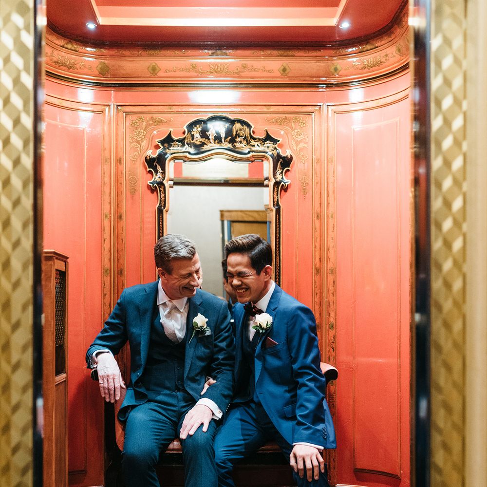 Grooms laugh together with orange in the background and gold gilded mirror
