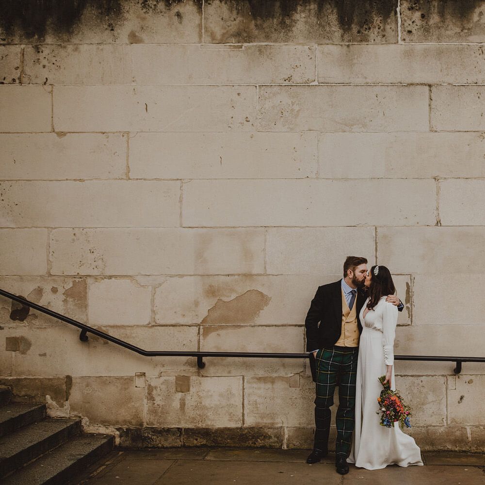 Bride and groom kissing against stone wall in London, bride wears long sleeve gown, groom wears three piece suit with tartan trousers 