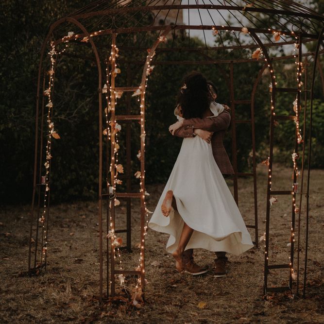 Groom picking up his bride in a wore framed pergola decorated with fairy lights 