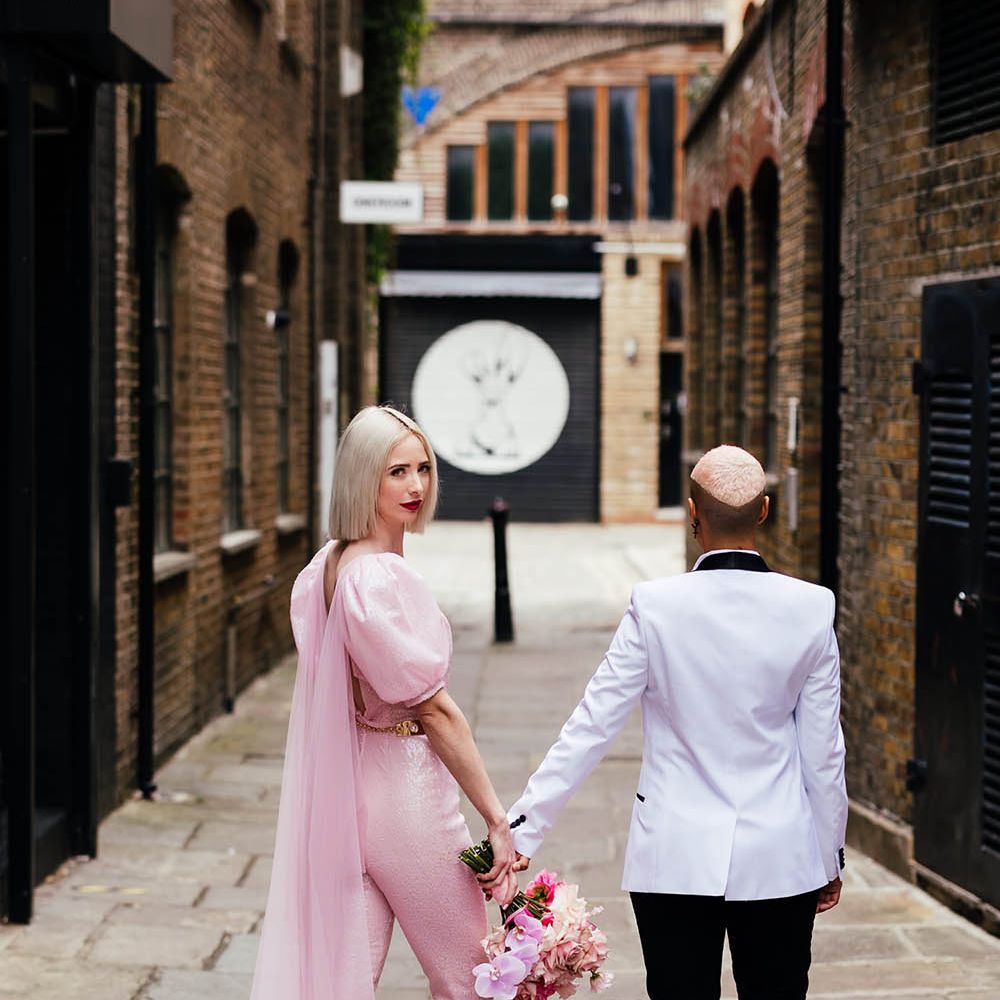Bride in a fitted pink jumpsuit with a Watteau train and pink mules holding hands with her bride in a white tuxedo 