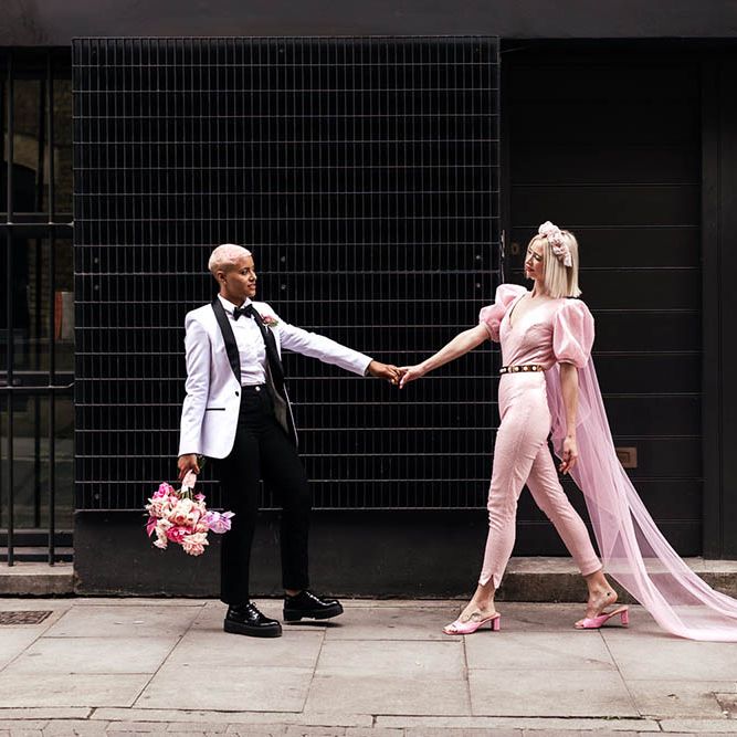 Bride in a white tuxedo jacket holding hands with her bride in a pink fitted jumpsuit with a Watteau train 