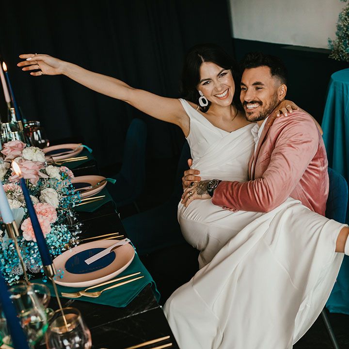 Bride in white boots with block heels sitting on her grooms lap at their pink and teal wedding reception 
