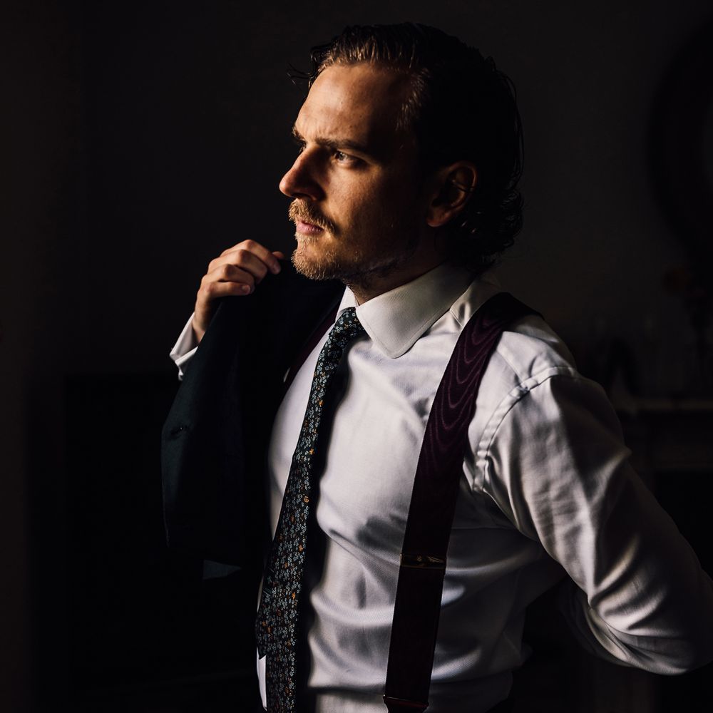 Groom in white shirt, patterned tie and braces puts on jacket by window for Cornish wedding with Halfpenny London Mayfair dress