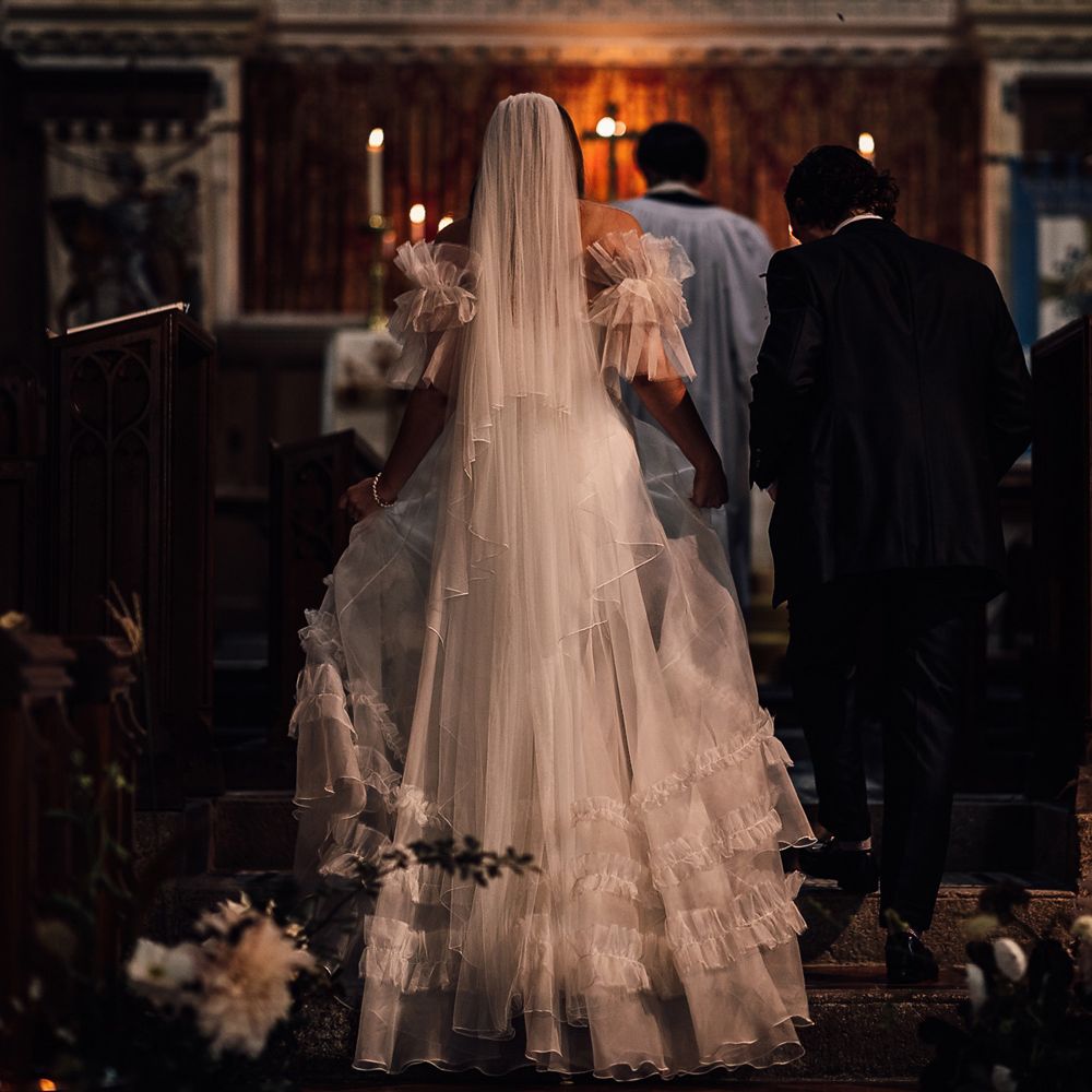 Bride in white Halfpenny London Mayfair dress with ruffle sleeves walks up to the altar with groom in suit during church wedding ceremony in Cornwall