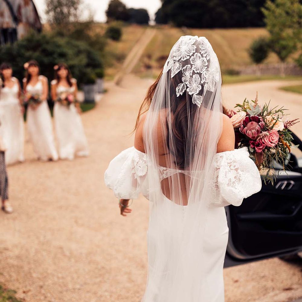 Bridesmaids see bride for the first time as she holds bright floral bouquet and wears lace embellished veil 