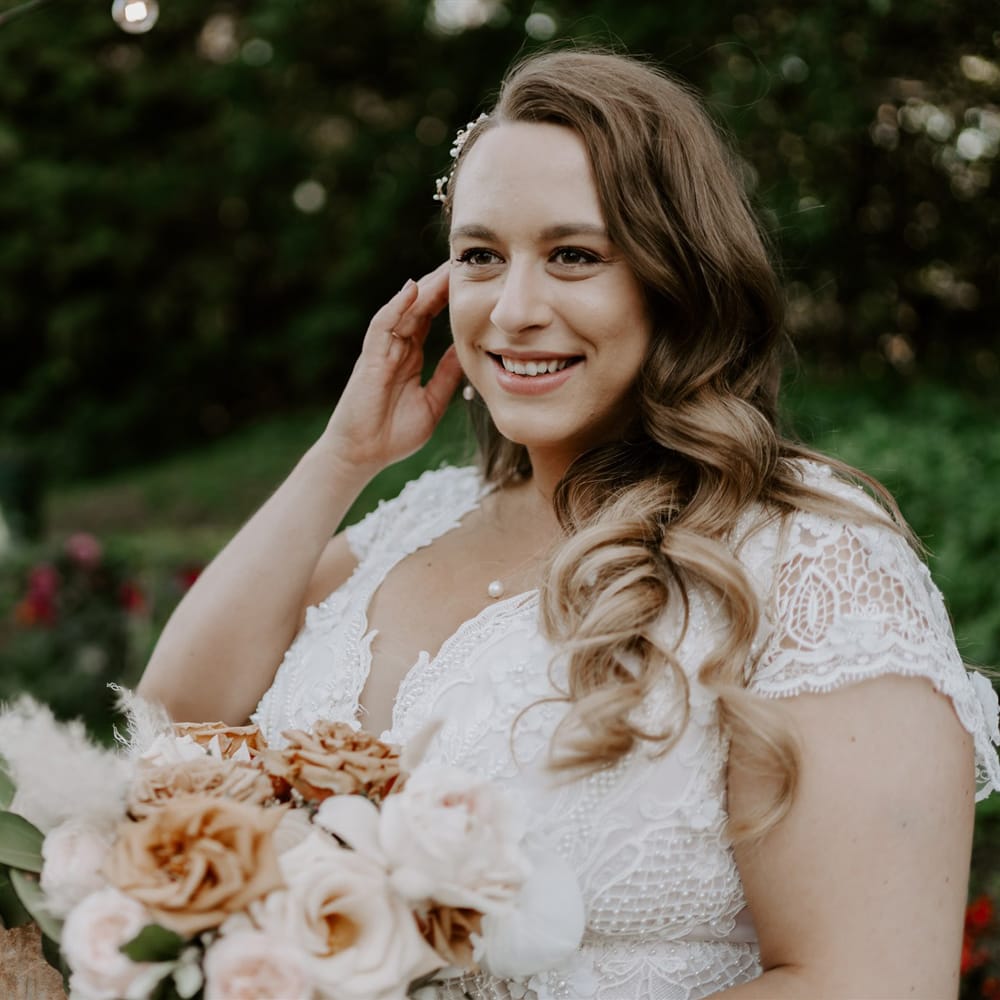 Smiling bride with long curled hair in homemade lace wedding dress and pearl necklace holds mixed rose bridal bouquet as she stands in garden after wedding ceremony