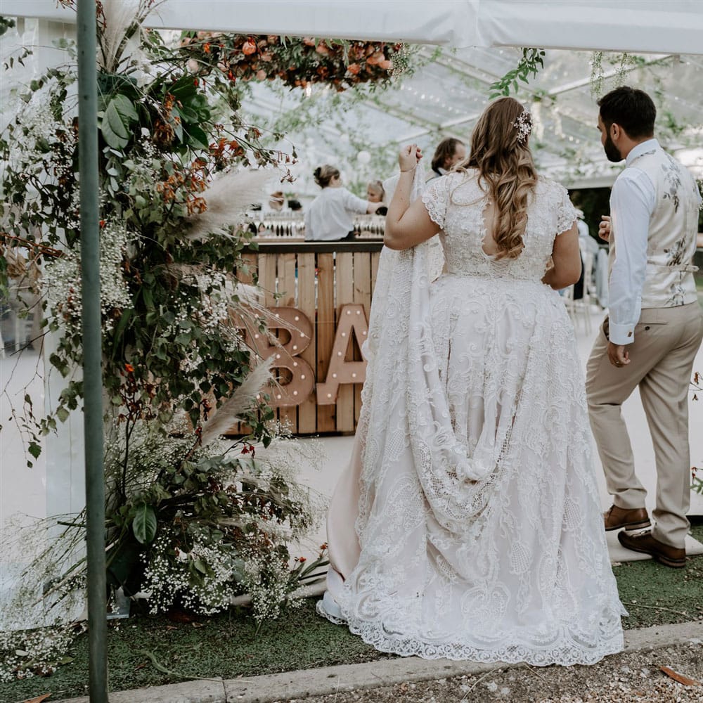 Bride in lace homemade wedding dress with train and open back with curled hair and groom in beige waistcoat and trousers walk into marquee with garlands at outdoor wedding reception
