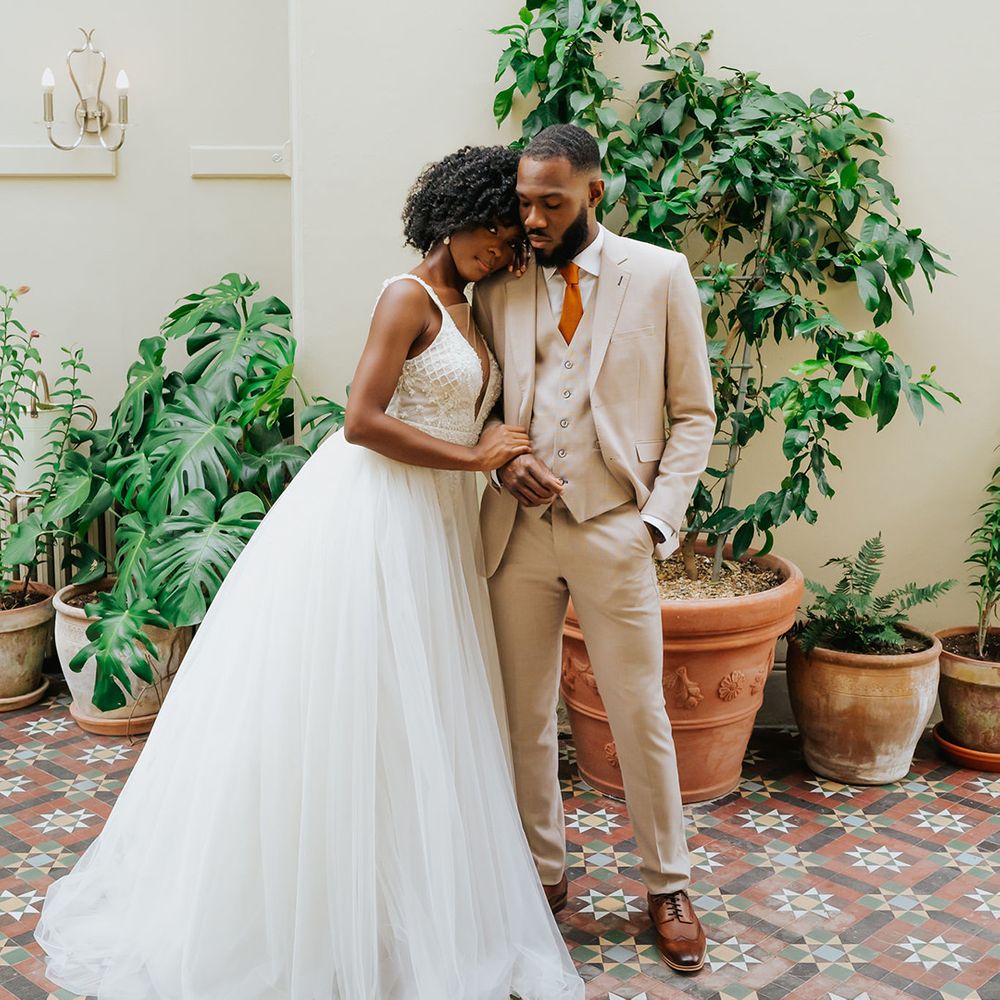 Black bride in a princess wedding dress with tulle skirt embracing her groom in a beige suit with orange tie in a conservatory at Modern Hall, London 