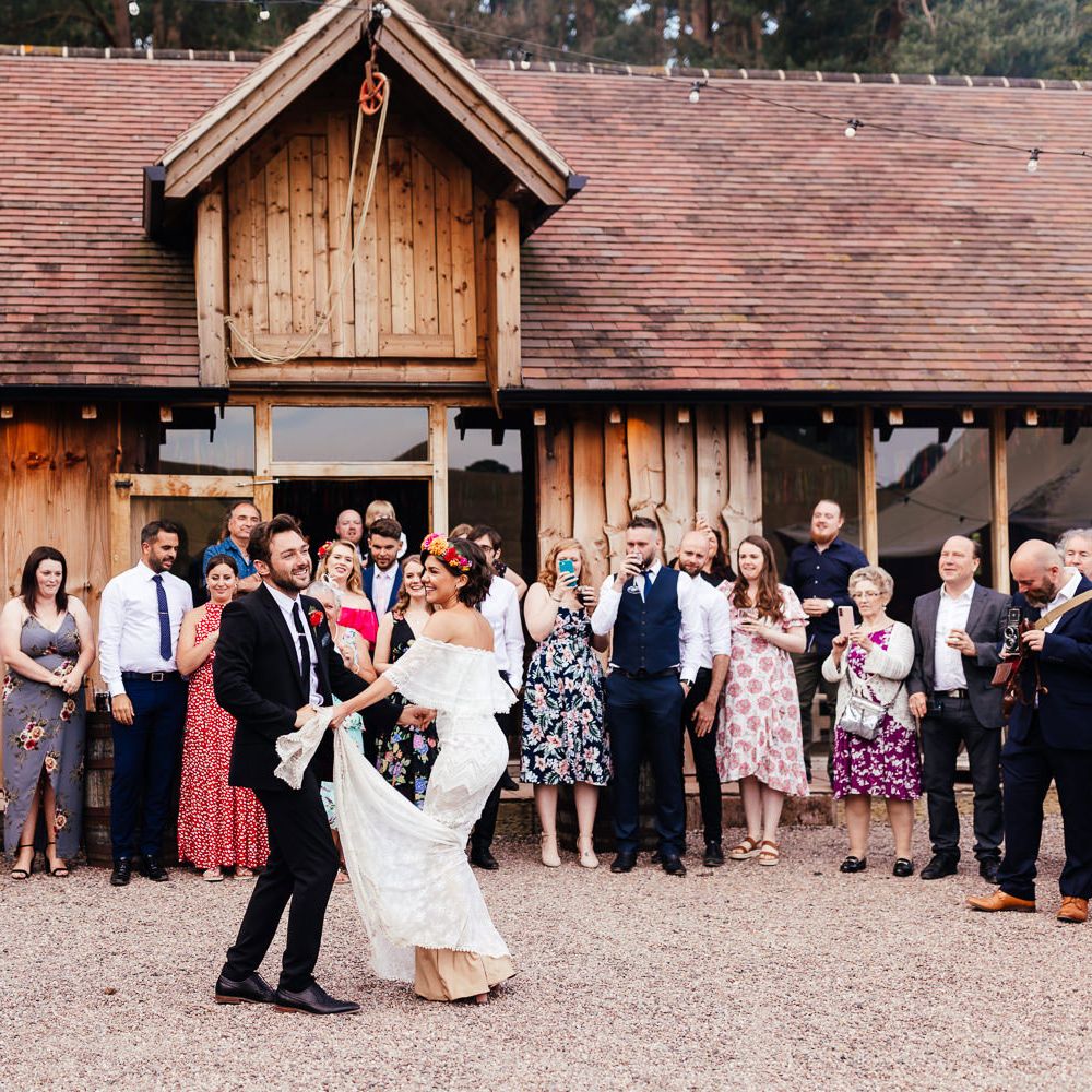 Bride in off the shoulder Grace Loves Lace wedding dress and flower crown dances with groom in black suit in front of wedding guests outside The Bridal Barn in Claverley