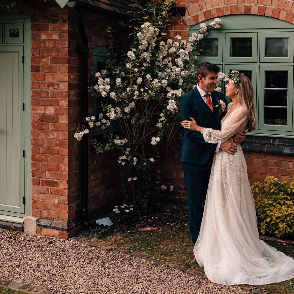 Groom in blue three piece checked suit hugs bride in long mesh sleeve Julie Vino wedding dress and flower crown as they stand outside cottage