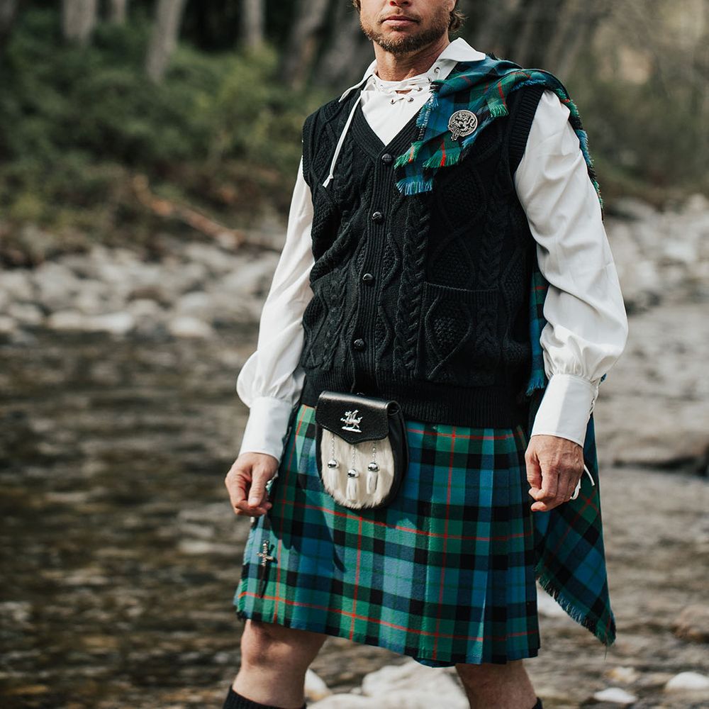 Bearded groom with long wavy hair in tartan kilt and  aran tank top walking through a stream 