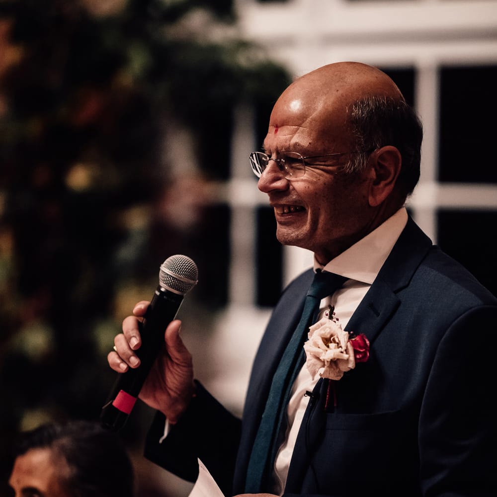Father of the bride in blue suit with floral buttonhole holds microphone as he makes speech at wedding reception