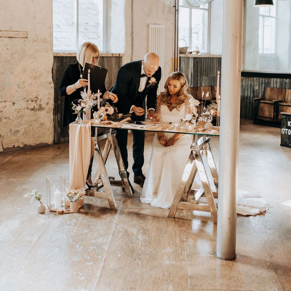 Bride in v neck wedding dress with sleeves sits signing wedding register with groom in navy tweed suit at Holmes Mill wedding