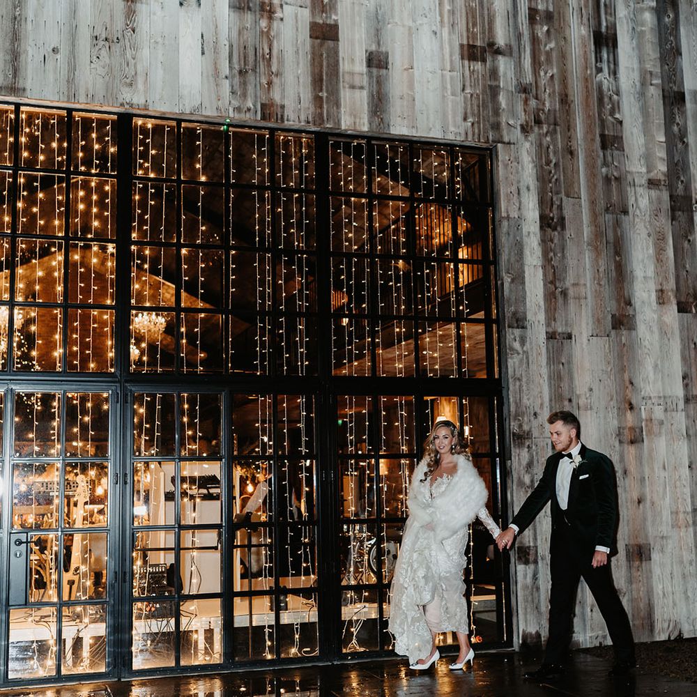 Bride in lace fitted wedding dress with white fur shawl walks with groom in black tie in front of fairy lights wedding
