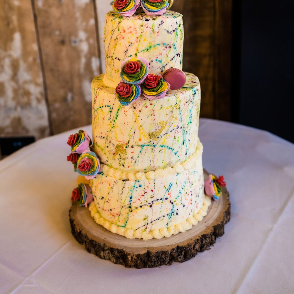 Three tier white frosted rainbow paint splattered wedding cake with rainbow roses, macarons and personalised wooden heart topper