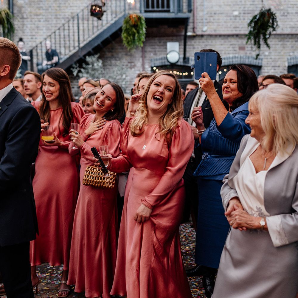 Bridal party in long sleeve pink satin bridesmaid dresses laughing during the wedding speeches 