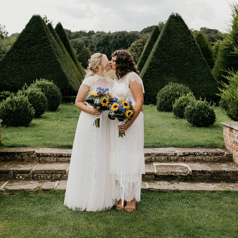 Brides holding sunflower and gypsophila wedding bouquets and sheer high neck wedding dresses share a kiss in the gardens of the venue