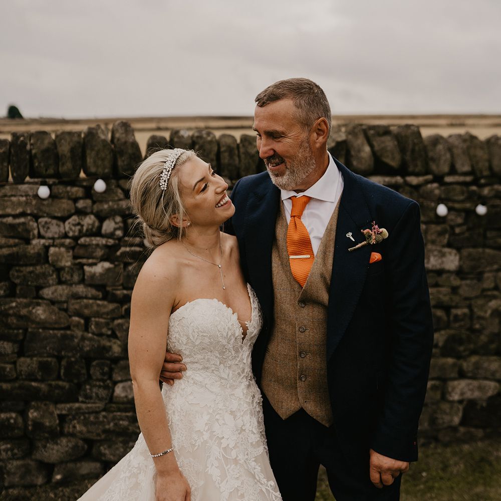 Father of the bride in dark suit jacket brown waistcoat and orange tie hugs the bride in sweetheart princess dress with tiara