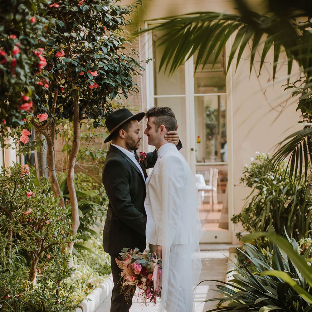 Groom in a black suit and fedora hat embracing his partner in a white groom suit 