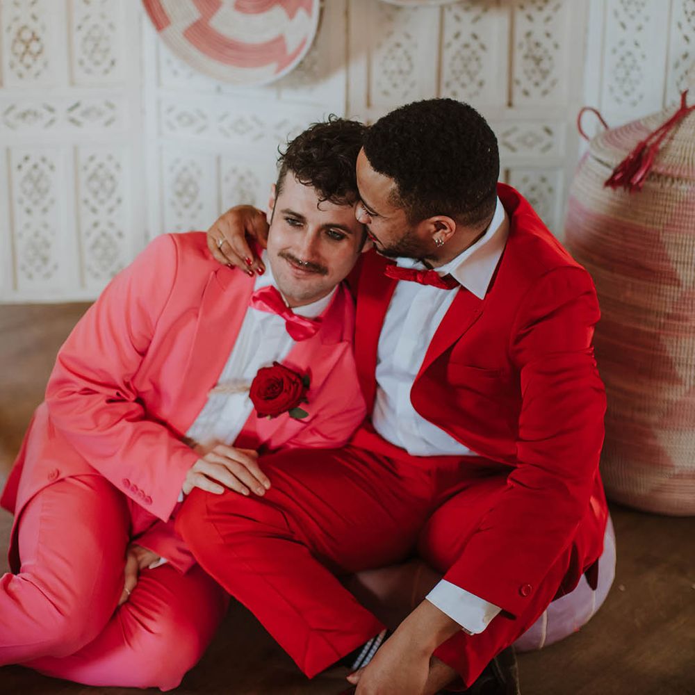 Groom in a red suit with red nail polish embracing his partner on the floor in front of a cream screen and wicker baskets in a pink suit 