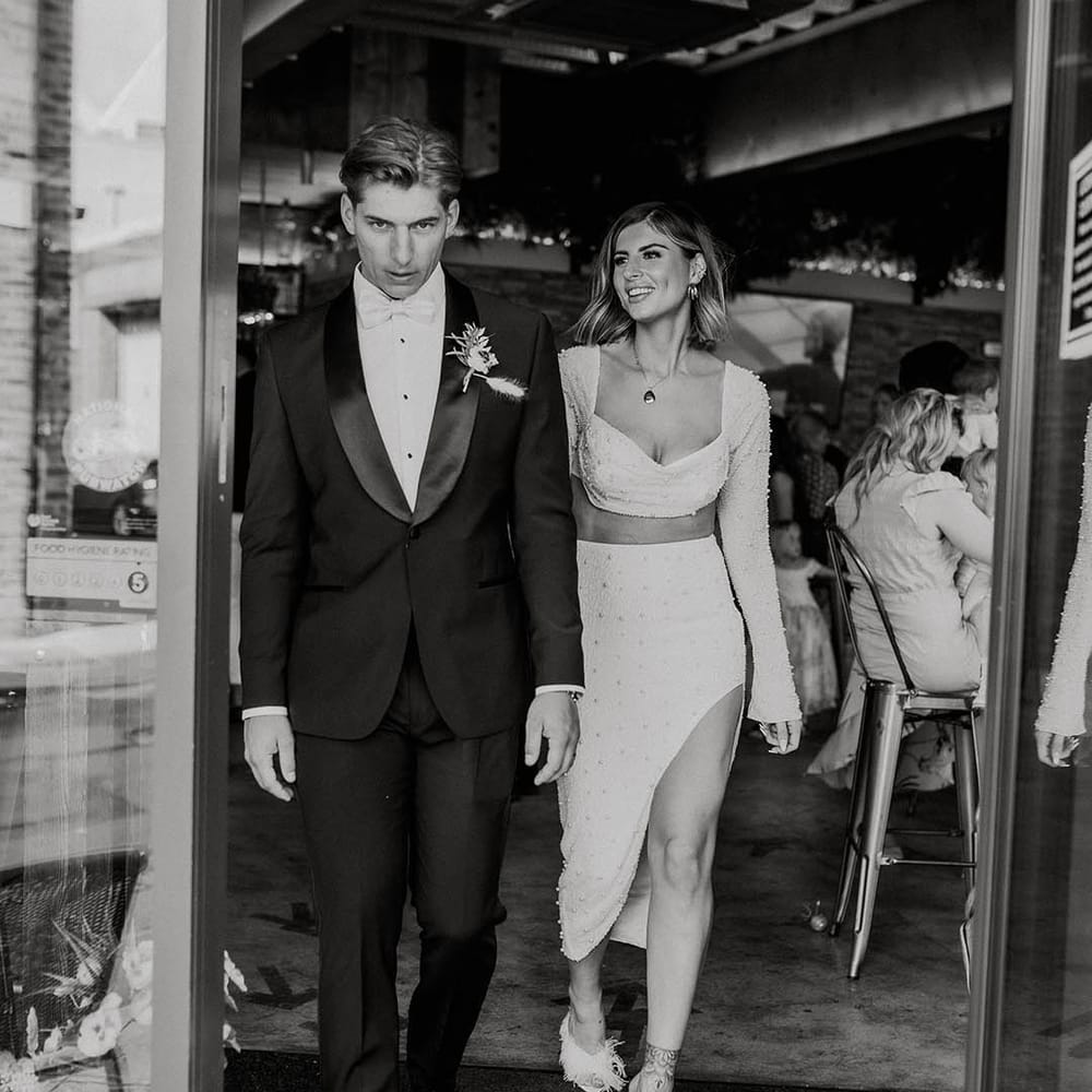 Groom in black suit jacket with satin lapels walking with bride in reception outfit two piece with pearls 