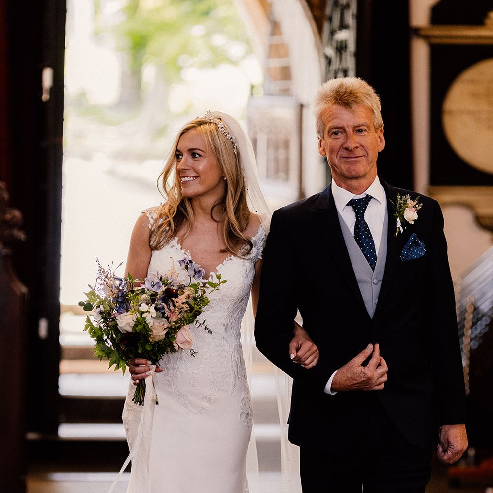 Bride with French tip nails and pearl headband wearing lace wedding dress is walked down the aisle by her father wearing a black and blue suit with polka dots