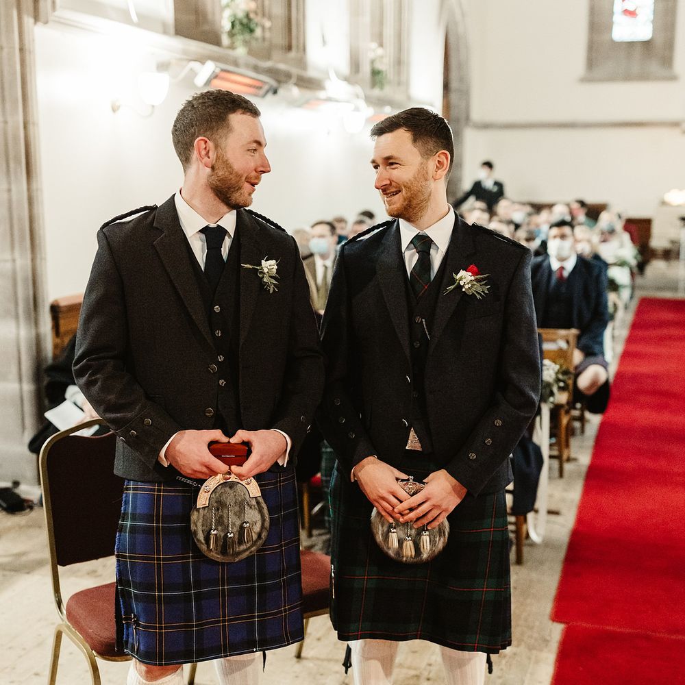 Groom awaits his bride with groomsmen at the front of church and both wear tartan kilts complete with white and red floral buttonholes 
