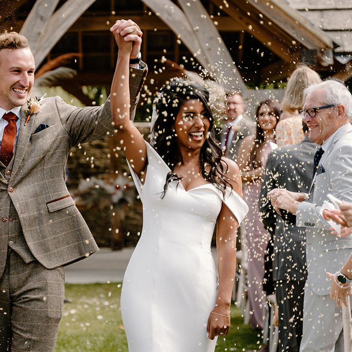 Groom in a brown suit with an orange tie raises his joint hands with the bride in a fitted wedding dress as they have a confetti moment 