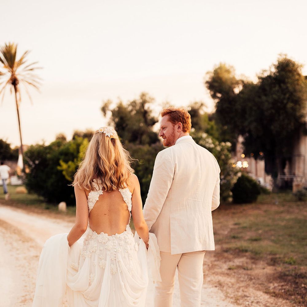 Bride in a flower halter neck wedding dress with flower hair accessories walking with the groom in a cream suit 