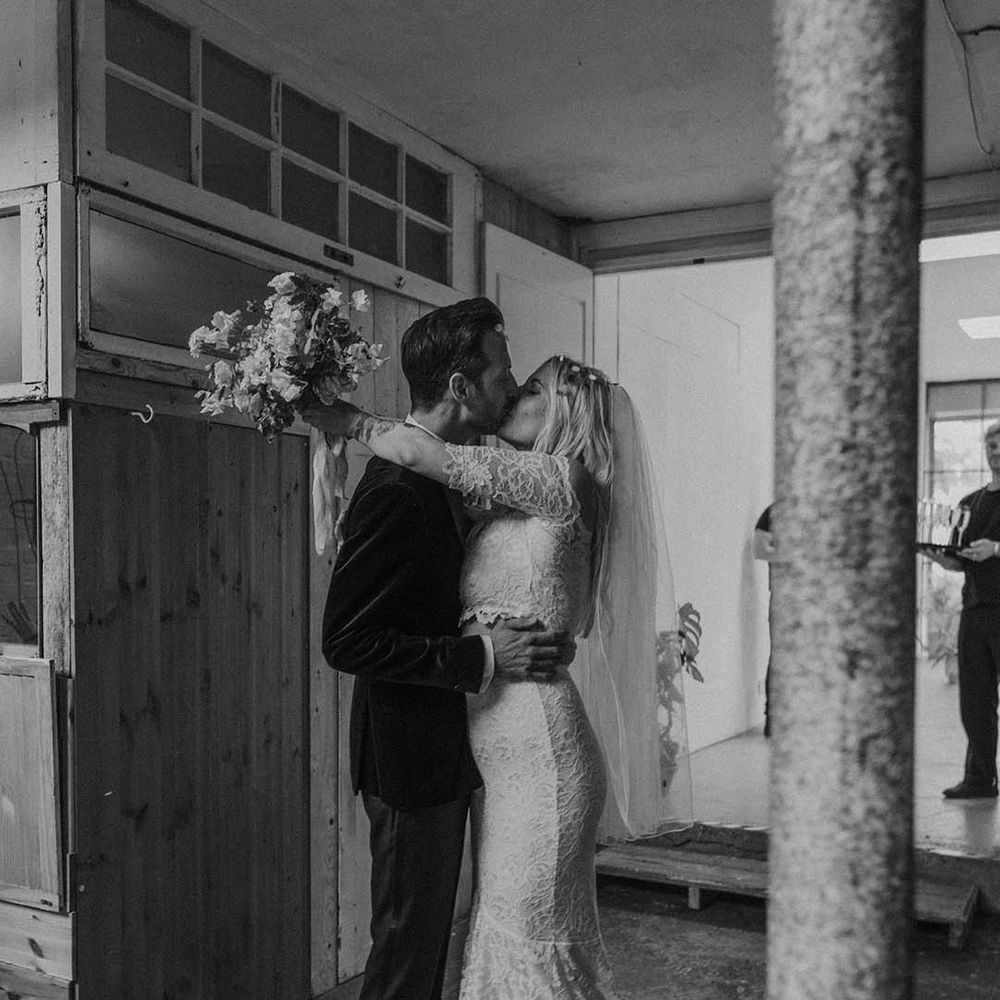 Bride in lace boho two piece with short veil and groom in black tux with bow tie and boutonniere kissing at Hackney Studios
