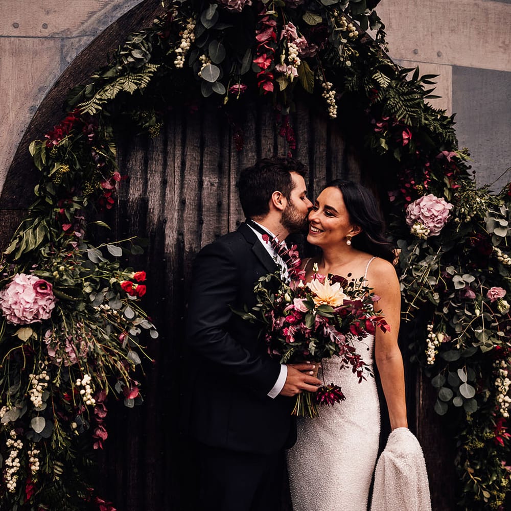 Groom in black tie kisses the bride on the cheek as they stand under a flower arch 