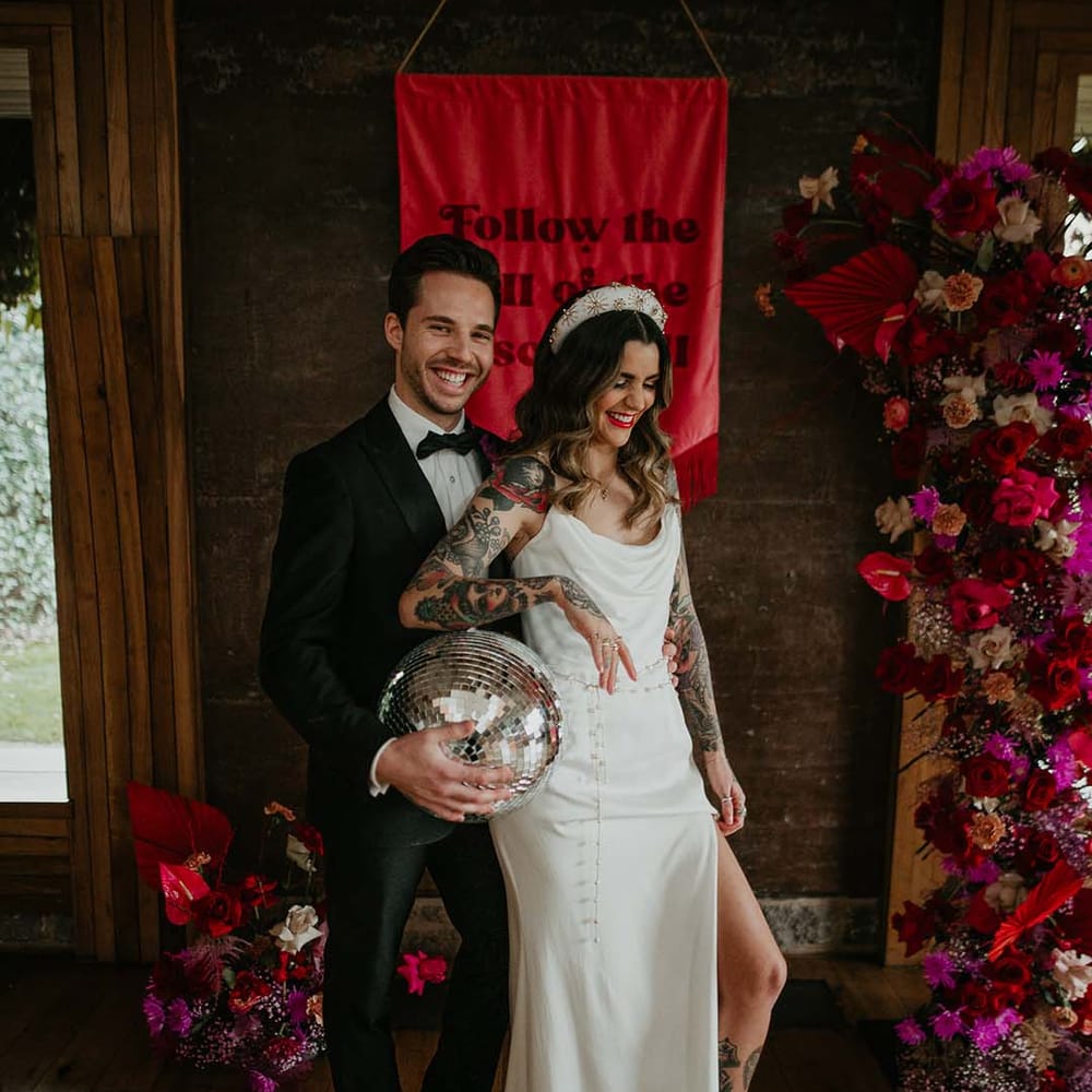 Bride in cowl neck sleeveless satin wedding dress with leg slit, gold glittery wedding headband, and soft grey fluffy feather open toe strappy high heels and groom in classic black tuxedo with magenta amaryllis and orchid boutonniere holding large disco ball wedding decoration with viva magenta fabric wedding banner and large wedding floral decorations 