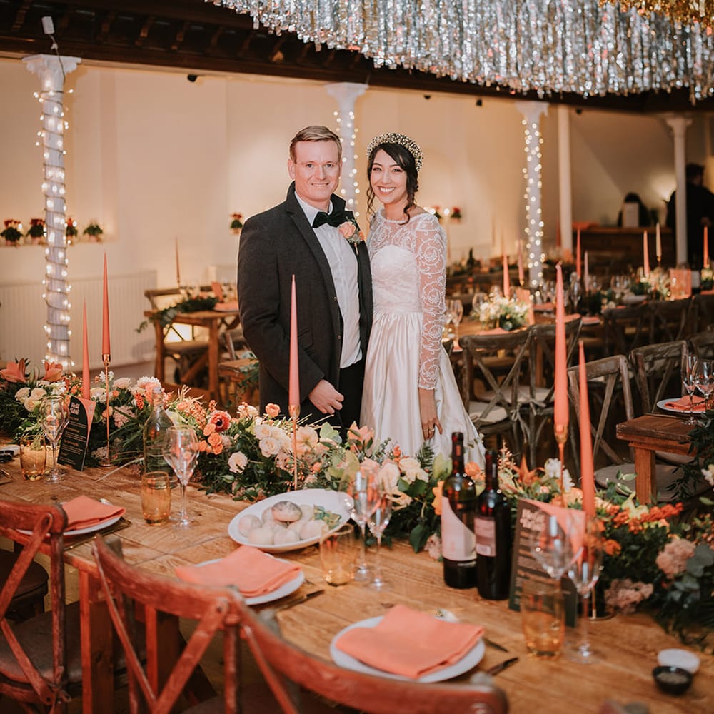 Bride and groom standing in reception room of Marylebone Town Hall with silver and gold wedding streamers, orange tapered candles, orange napkins, orange rose and foliage tablerunners and fairy lights 