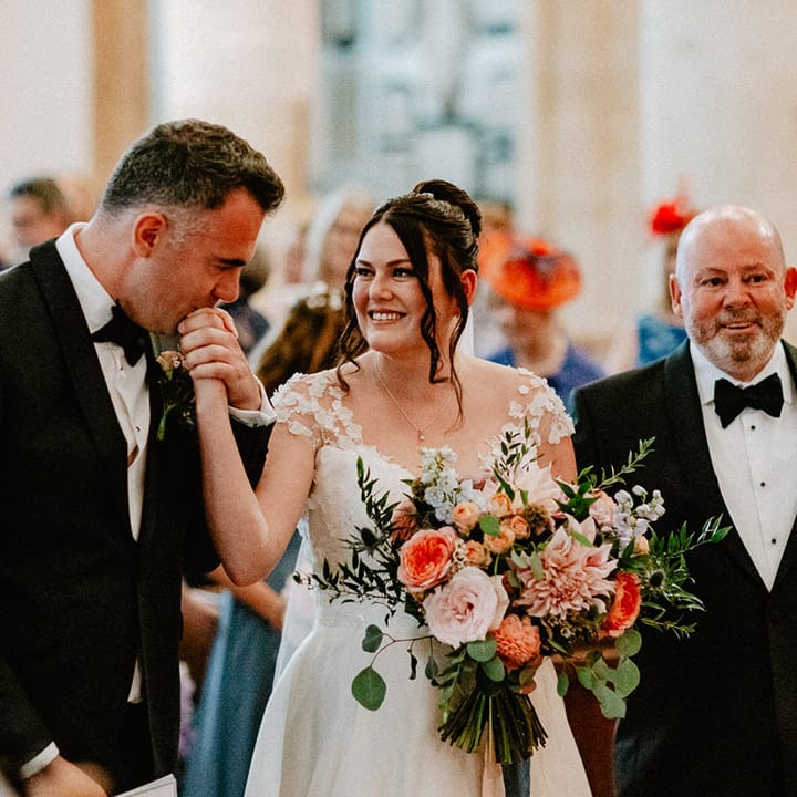 Groom wearing a black tuxedo kisses the bride's hand as she walks down the aisle to meet him for the start of the church wedding ceremony 
