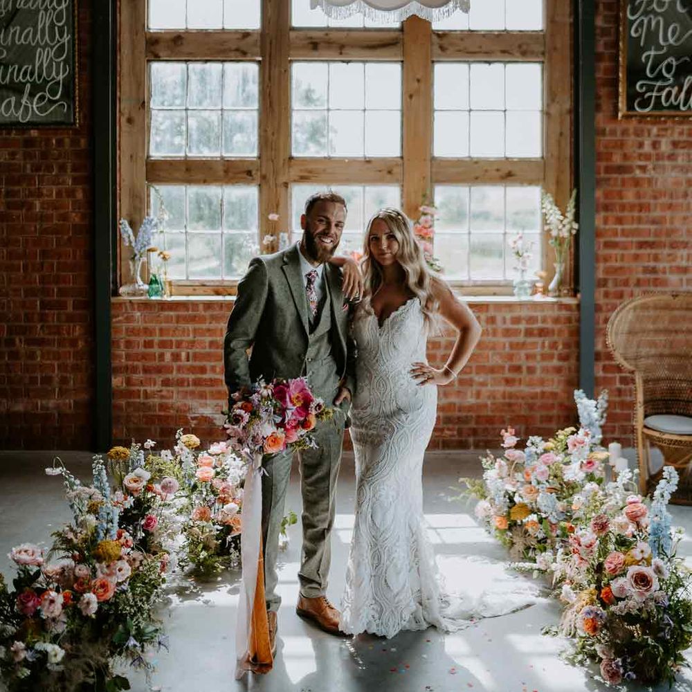 Bride in v neck lace wedding dress and groom in grey three piece suit standing under fabric lamp shades surrounded by colourful flowers at The Giraffe Shed 