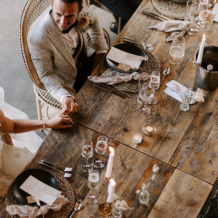 The bride and groom sit on boho peacock chairs for their wedding breakfast at wooden banquet tables with wicker placemats and black plates 