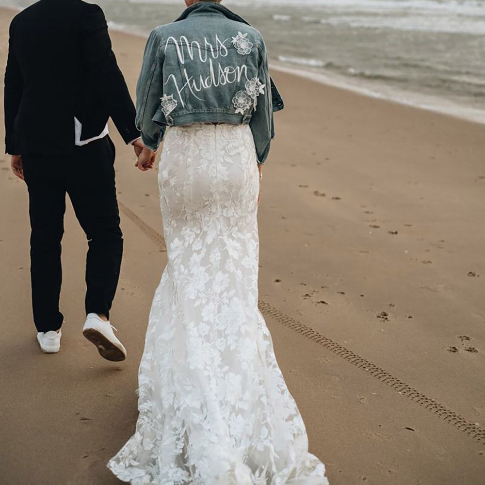 Bride in lace wedding dress wearing a denim bridal cover-up walking on the beach with the groom with hand painted new last name and 3D flowers