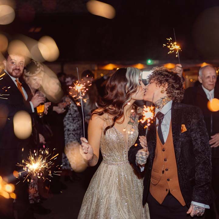 Bride in dark velvet bridal jacket with rust waistcoat and pocket square, black suit trousers and gold pocket watch kissing bride in sleeveless deep v neck sparkly gold wedding dress doing wedding sparkler send off at Kingscote Barn Tetbury LGBTQIA+ wedding