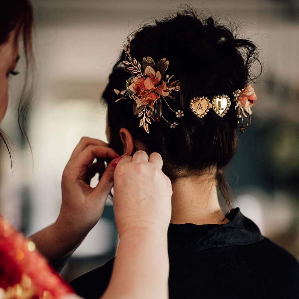 Bride with dark brown wedding hair being secured into an updo with flower hair accessories and personalised gold and pearl heart hair pins 