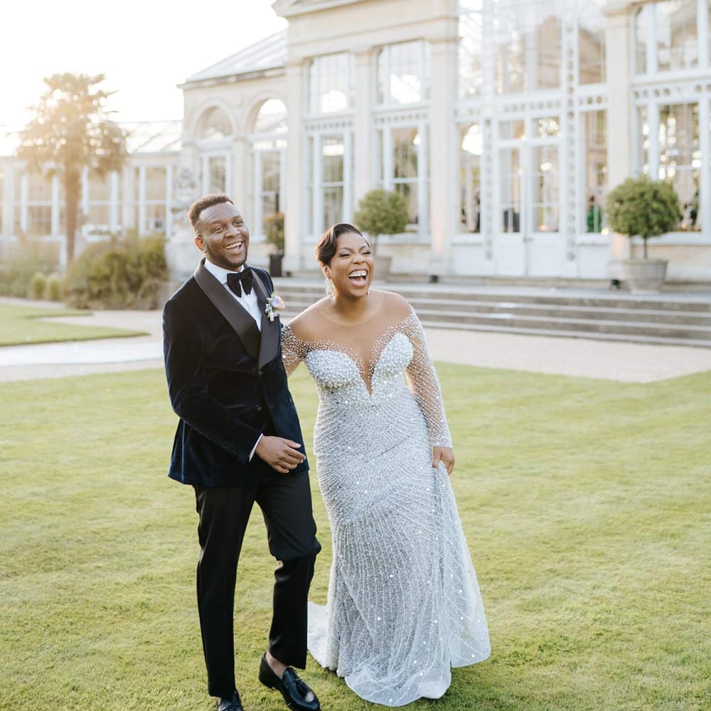 Bride in illusion v neck sheer off shoulder long sleeve pearl embellished wedding gown laughing with groom in velvet midnight blue double breasted groom’s blazer, velvet bowtie and peach garden rose boutonniere during vow renewal at Syon Park wedding venue
