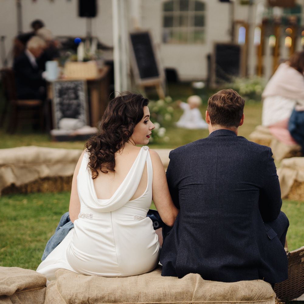 Bride & groom sit on hay bale at picnic 