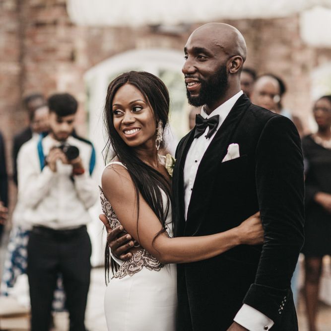 Black Bride with long hair embracing her husband in a velvet tuxedo jacket on the dance floor 