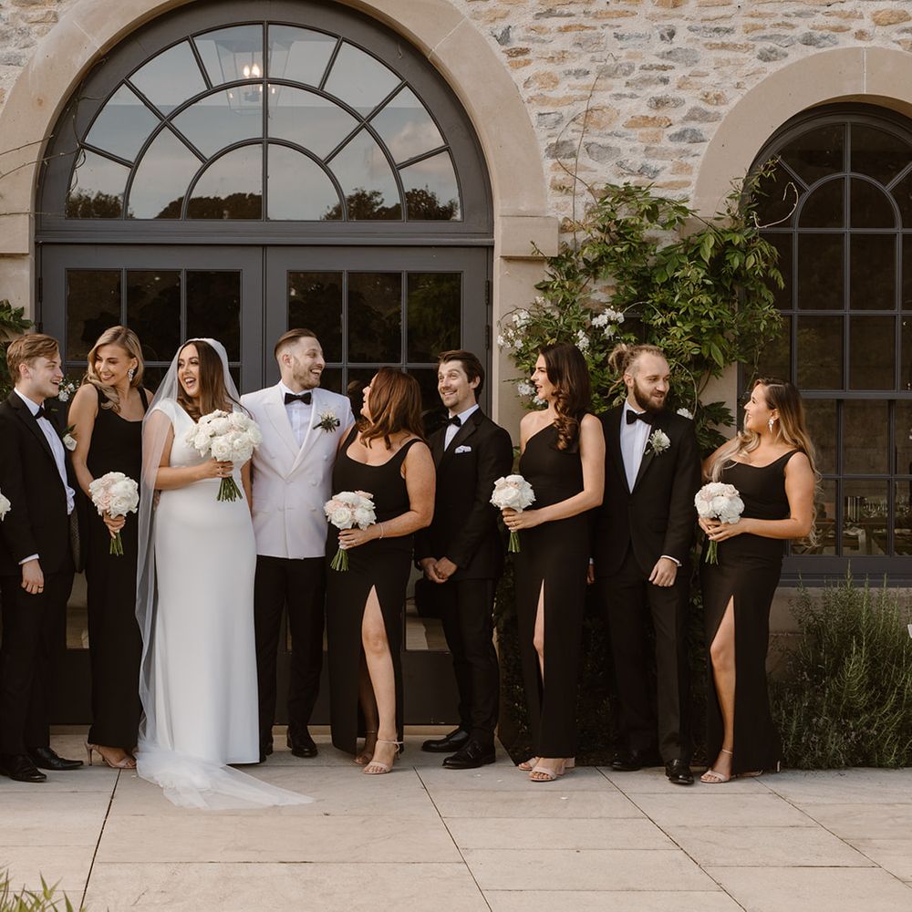 Bride in white Made With Love wedding dress poses outside with groom in white tuxedo jacket, bridesmaids in long black bridesmaid dresses and groomsmen in black tuxedos