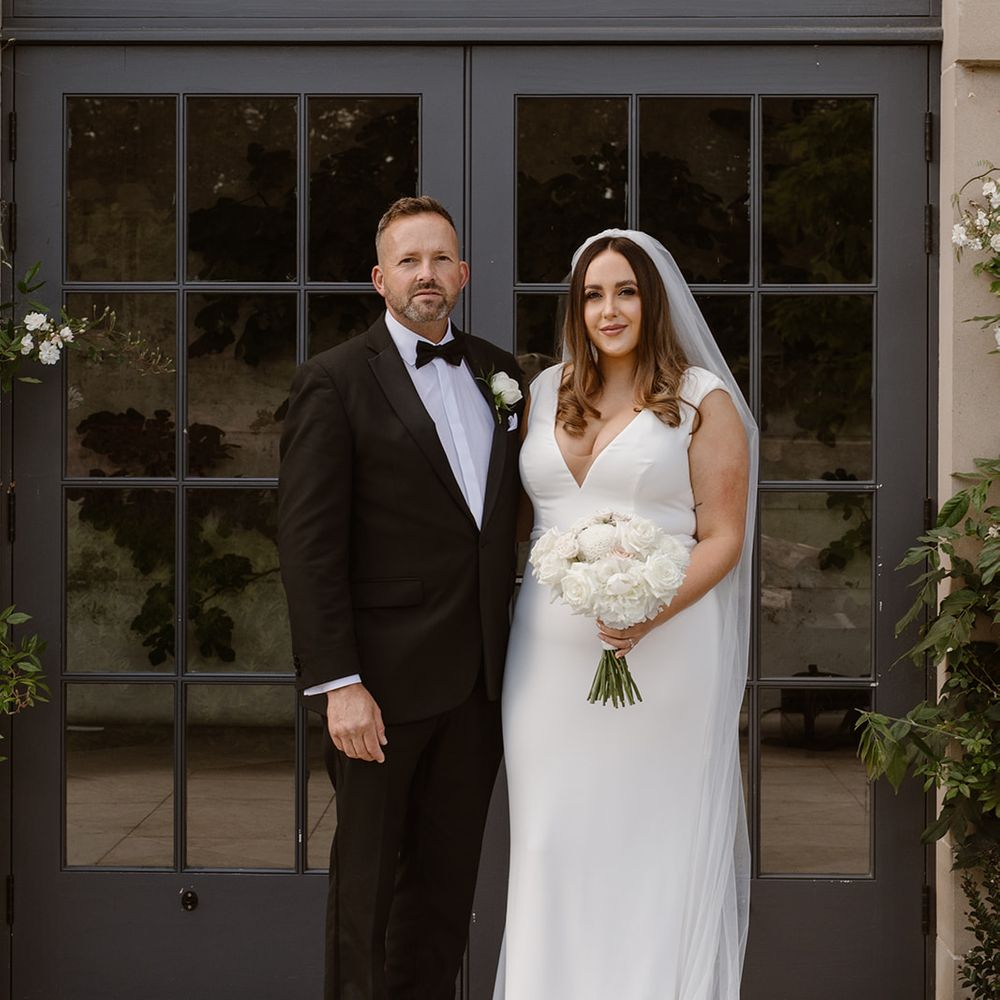 Bride in white Made With Love wedding dress holding white and blush pink rose bouquet poses outside with father in black tuxedo