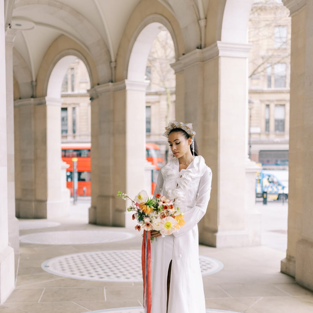 Bride wears golden crown with ruffled front wedding gown whilst holding bright and bold floral bouquet tied with pastel velvet ribbon
