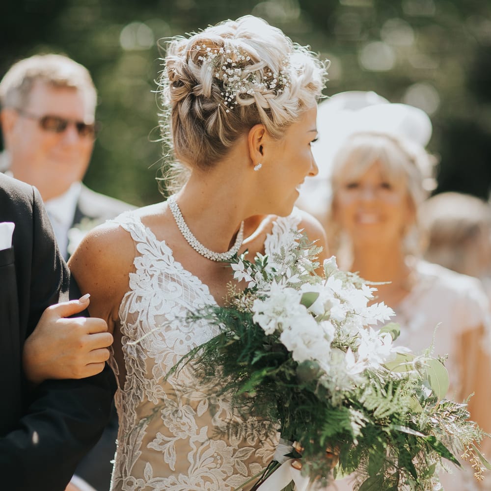 A bride in a lace wedding dress carries a bouquet as she walks down an outdoor aisle. Her blonde hair is up high with a braid and gypsophelia in the side.